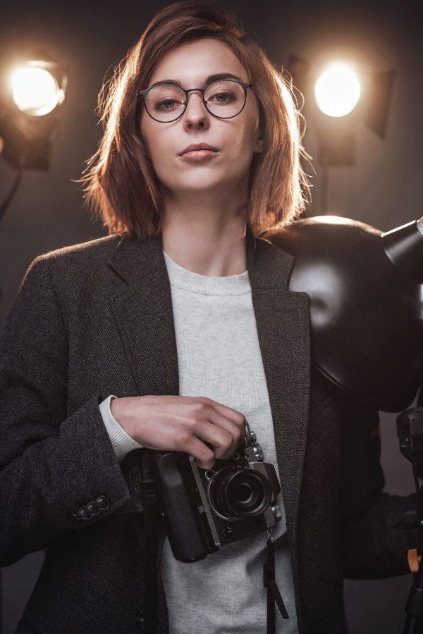 Portrait of a beautiful redhead female photographer holds a digital camera and looking on the camera. Studio shot on dark background with lighting equipment